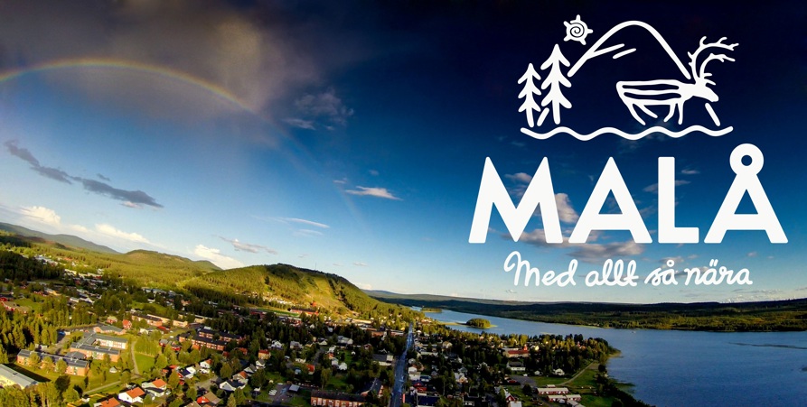 Flygbild över Malå samhälle. Regnbåge över berg och skog i fjärran. Malåns vatten. 