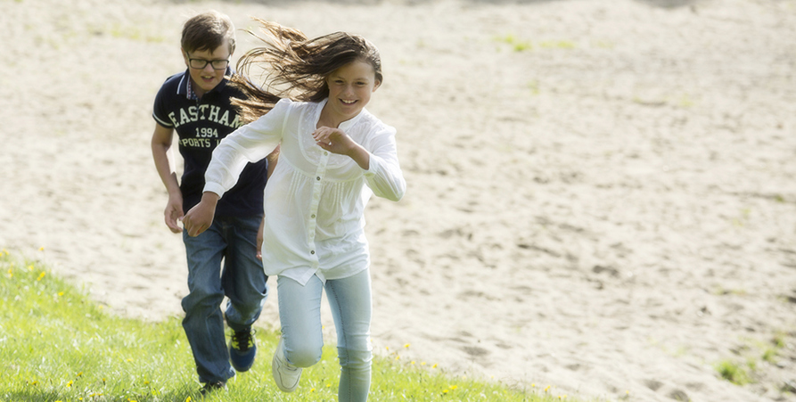 Två ungdomar springer uppför en grässlänt med badstrand i bakgrunden, sommar. 
